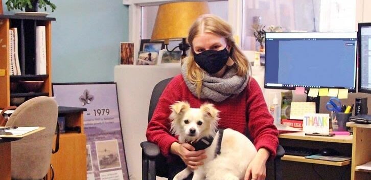 Blonde woman wearing a face mask sits at a desk in an office with a small white dog on her lap